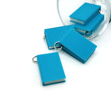 A 3D miniature book charm in teal-blue, approx. 27×18 mm, with a textured cover and a jump ring attached through a 3 mm hole—displayed on a white background.