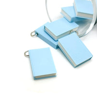 A miniature 3D light blue book charm, approximately 27×18 mm, with a textured cover and attached jump ring, displayed against a clean white background.