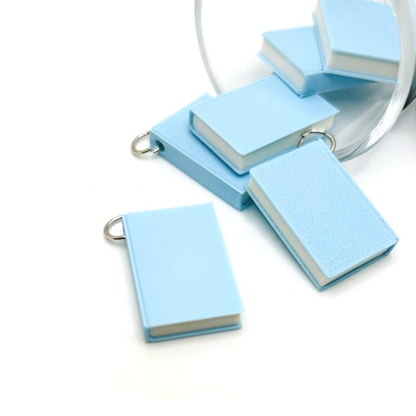 A miniature 3D light blue book charm, approximately 27×18 mm, with a textured cover and attached jump ring, displayed against a clean white background.